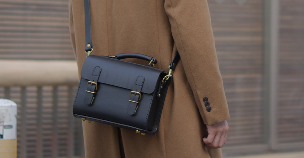 黑色皮革迷你單肩包，設有金色搭扣和可調式肩帶，包身俐落，展現極簡風格。