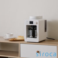 白色 Siroca SC-A3520 自動研磨咖啡機，配有玻璃咖啡壺、黑色手柄，頂部有白色蓋子。咖啡機正面有黑色顯示螢幕和按鈕，左側有一組白色杯子和木質托盤，上面放著司康餅和餅乾。