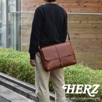 一個男人背著一個棕色皮革馬鞍包，包的正面有細長的皮帶和釦環，旁邊有綠色植物，包的右下角有 HERZ 的 Logo 和 HAND MADE IN JAPAN Since1973 的文字。
