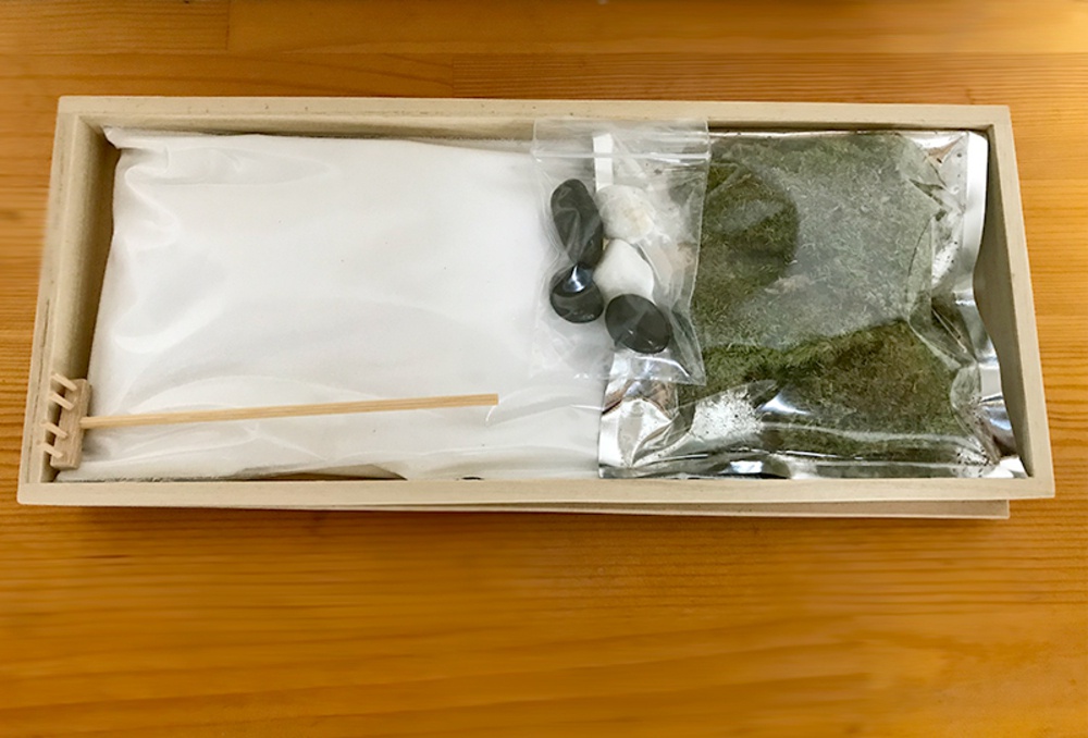 淺木盒內裝有日本枯山水模型組件，包含白色細沙、綠色苔蘚、黑色與白色小石子，以及一個木製耙子和一根竹籤。