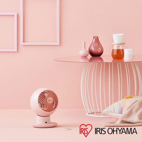 簡約的粉紅色圓形空氣循環扇，帶有可調節底座，擺放在粉色背景的桌面上，旁邊有裝飾性花瓶、茶壺、杯子和抱枕。