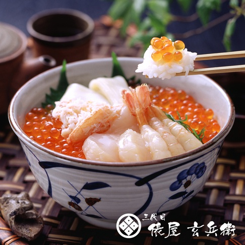 日式海鮮丼，碗內盛有白米飯，鋪滿鮭魚卵、鮮蝦、干貝及蟹肉。碗身有藍色花紋，背景為木質餐墊與茶具。