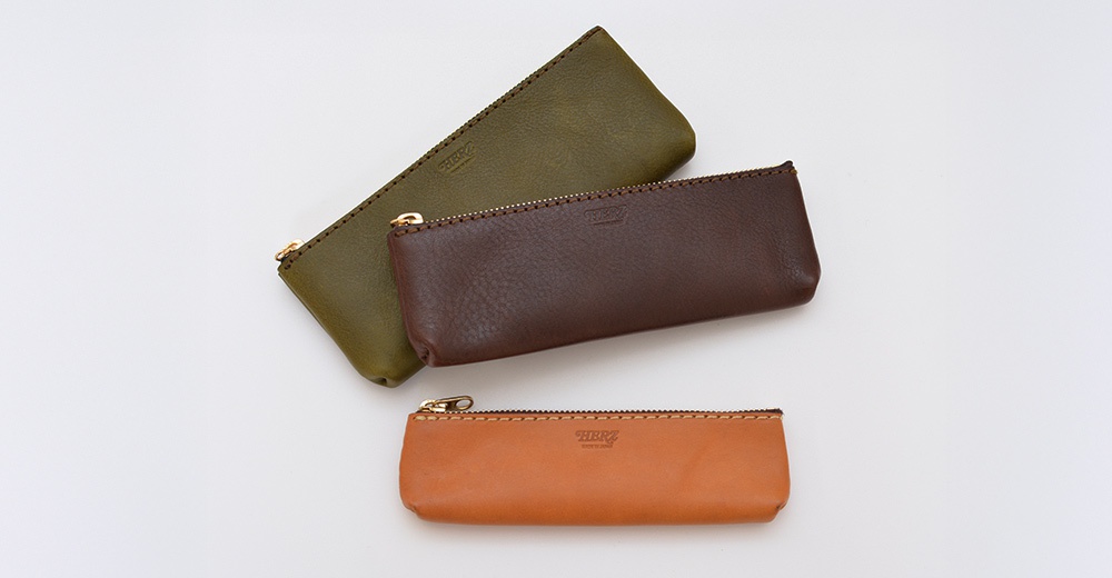 職人手作皮革收納筆袋，展示橄欖綠、深棕色及焦糖色三款，皆為拉鍊開合設計，材質為皮革，表面帶有細緻縫線。