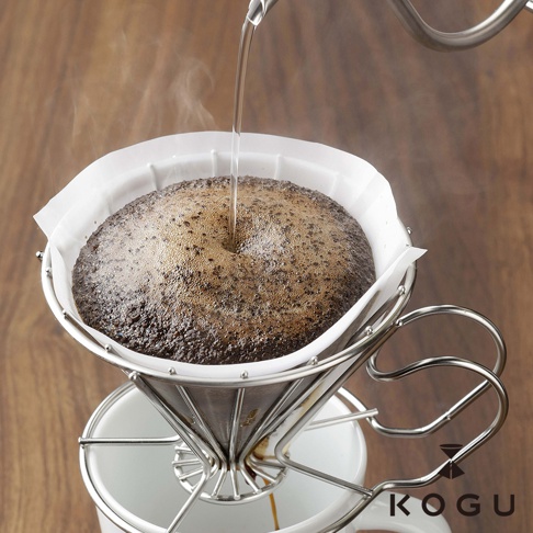 日本製銀色金屬濾杯，內含白色濾紙，正進行手沖咖啡，咖啡粉與熱水混合產生泡沫，濾杯架於白色馬克杯上。