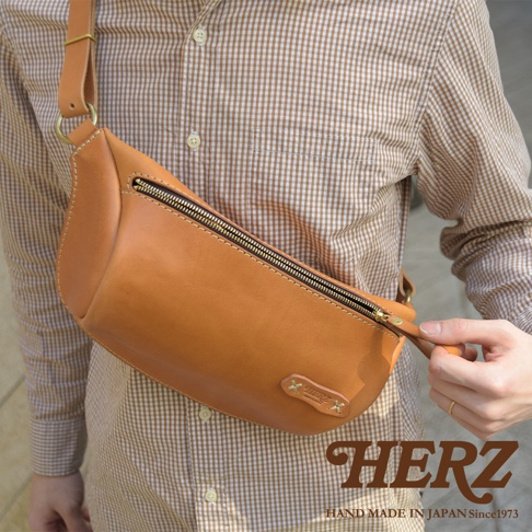 職人手作皮革輕便斜背包，採用鞣製皮革材質，呈現溫潤的天然棕色調。包款擁有弧線造型，搭配黃銅色拉鍊與車縫線細節，展現質感。