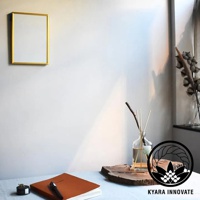 KYARA INNOVATE 品牌標誌旁為室內擴香竹，前方有一本橘色筆記本與筆，背景為留有日光陰影的白色牆面，牆上懸掛金色邊框相框，右側擺放著乾燥花束。