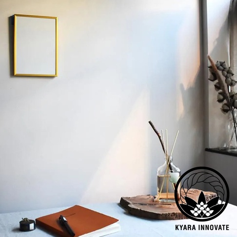KYARA INNOVATE 品牌標誌旁為室內擴香竹，前方有一本橘色筆記本與筆，背景為留有日光陰影的白色牆面，牆上懸掛金色邊框相框，右側擺放著乾燥花束。