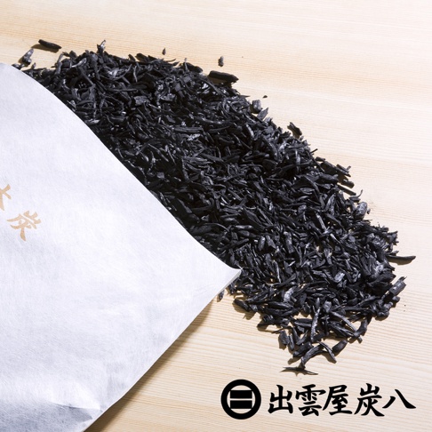 一堆細碎的黑色木炭散落在淺色木紋背景上，部分木炭從白色紙袋中傾倒而出，紙袋上有金色日文字樣。