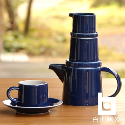 藍色陶瓷 M 型壺與同色系的咖啡杯及盤，壺身上有層疊設計，杯口及壺身邊緣為白色。