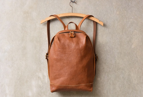 棕色皮革後背包，簡約褶皺設計，雙拉鍊開口，皮革肩帶，掛於木質衣架上，背景為淺灰色紋理牆面。