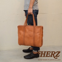 一只手提著棕色皮革托特包，包的頂部有拉鍊開合，正面有一個小口袋。