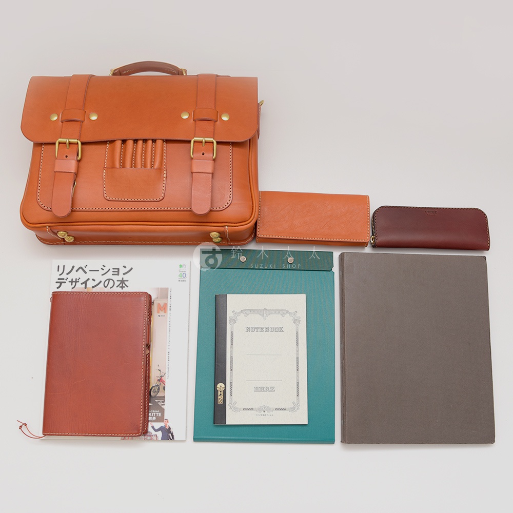 職人手作皮製歐風三用學生包，配有黃銅釦環和皮帶。旁邊擺放著同色系的長夾、一個拉鍊筆袋，以及一本綠色封面的筆記本和一本灰色筆記本。