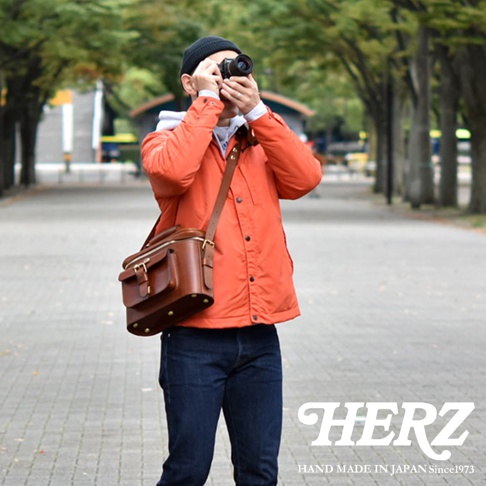 一名男子身穿橘色外套，戴著黑色毛帽，正在專注地用相機拍照。他身上揹著一個棕色的皮革相機包，包包上有金屬扣和皮帶。背景是秋天的公園，有著茂密的樹林和一條鋪著磚塊的小徑。圖片右下角有「HERZ」和「HAND MADE IN JAPAN Since1973」的文字。