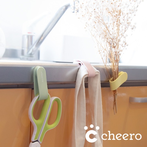 綠色萬能QQ變形夾夾著一把綠白相間的剪刀，掛在廚房檯面上。旁邊有粉色和黃色變形夾，分別掛著白色毛巾和一束乾燥花。
