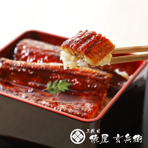 紅褐色醬汁烤鰻魚覆蓋在白米飯上，盛裝在黑紅相間的漆器盒中，一雙筷子夾起一塊帶有烤紋路的鰻魚和米飯，背景為白色，右下方有日文品牌標誌。
