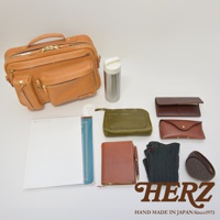 職人手作皮革兩用單肩包，呈現天然皮革的淺棕色，包身設計多個拉鍊口袋和翻蓋口袋。周邊配件包含軍綠色皮革小包、棕色長夾、棕色眼鏡盒、筆記本、鋼筆、灰色襪子與棕色毛絨耳罩。