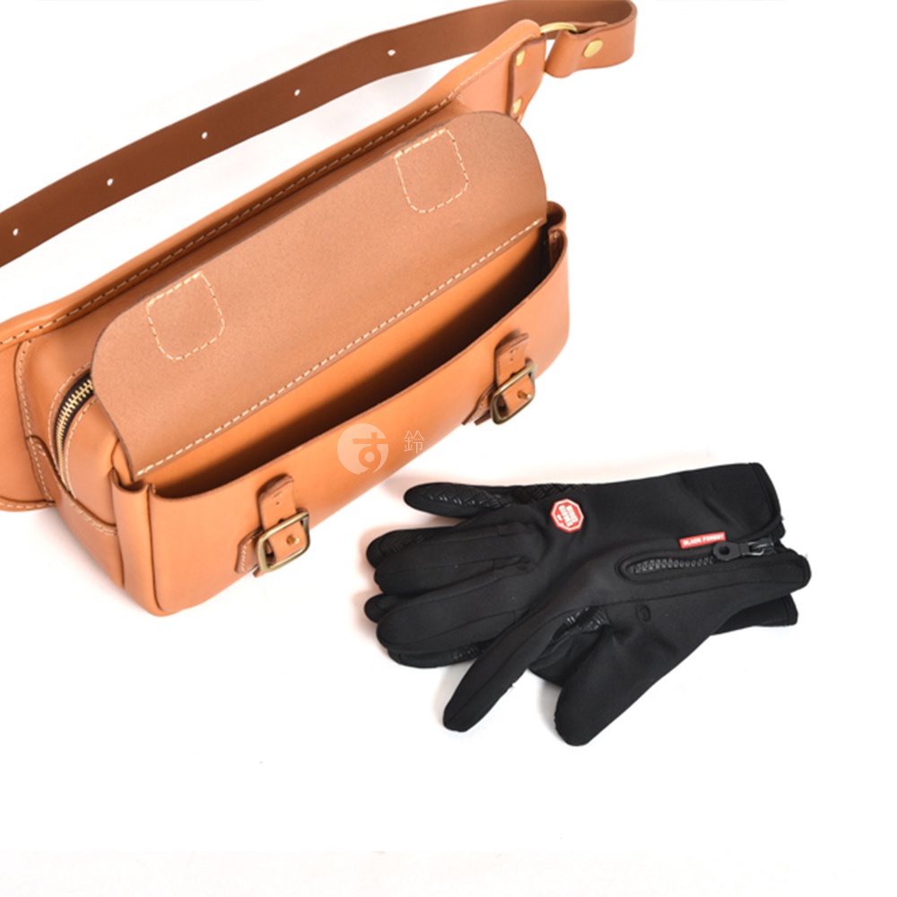 棕色皮革騎士斜背包，帶有翻蓋和兩個金屬扣，旁邊是一副黑色防風手套。