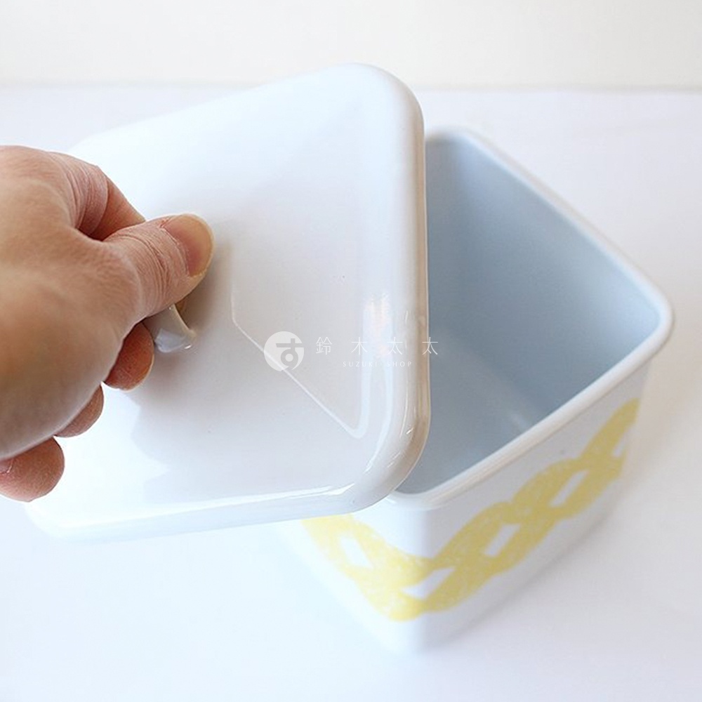 一只手正在拿著一個白色琺瑯方形收納盒的蓋子，盒身有黃色編織狀圖案。