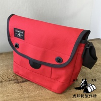 紅色純綿帆布雙扣郵差包，配有黑色滾邊、黑色扣子和黑色肩帶。包的正面有一個黑色皮革裝飾片，上面有一個黑犬印製作品所的黑色標籤。