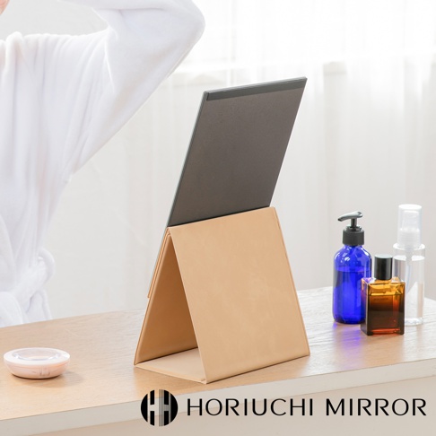 NAPURE 桌上型2Way折疊化妝鏡，米白色皮革外殼，鏡面為深灰色，可站立於桌面，搭配藍色壓頭瓶、香水瓶和噴霧瓶。