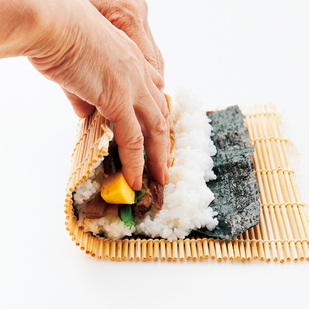 雙手正在竹簾上製作壽司，其中包含米飯、深綠色海苔和黃色、棕色的餡料。