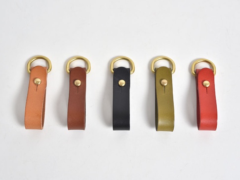 五個職人手作皮革吊掛扣帶，顏色分別為淺棕、深棕、黑色、橄欖綠、紅色，均採用真皮材質，搭配黃銅色D型環和鉚釘，簡約實用。