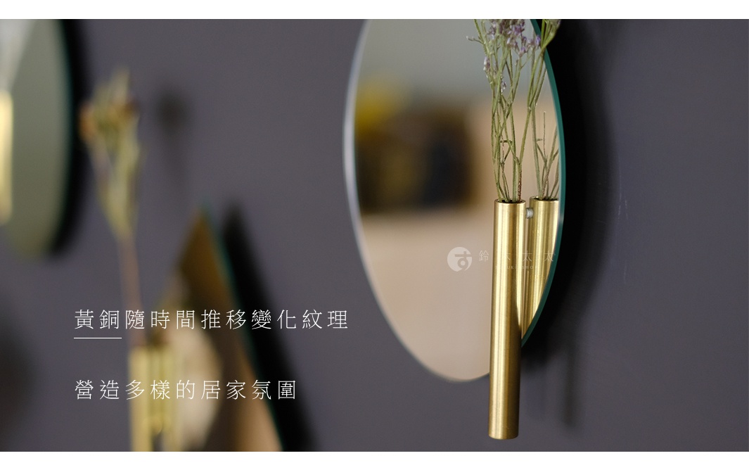 壁掛鏡面漂浮一輪花器，黃銅材質，設有兩個直立的細長圓筒，分別插入乾燥的細長綠葉植物，鏡面呈圓弧形，反射出溫暖的光澤。