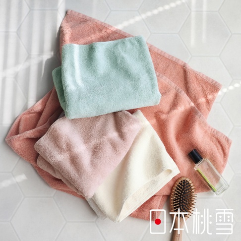 這是一張俯視視角的居家生活照片，背景為白色六角形磁磚，擺放了數條不同顏色的日本桃雪精梳棉毛巾，包含粉紅色、淺綠色與米白色，毛巾質地柔軟且蓬鬆。畫面右下方放置了一把木質梳子與一小瓶透明護理油，右下角印有日本桃雪品牌名稱。
