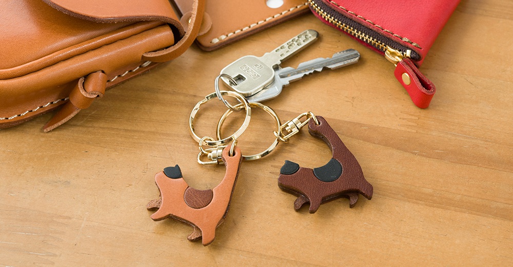皮革製貓咪造型鑰匙圈，一個為淺棕色，另一個為深棕色，均有黑色點綴，搭配金色鑰匙圈及鉤環。