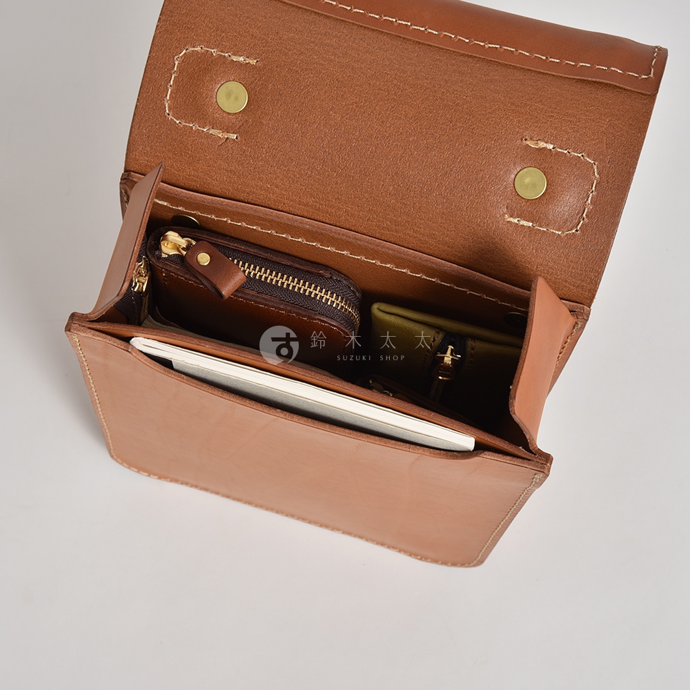 職人手作皮革立體口袋兩用單肩包，淺棕色皮革材質，包內有深棕色拉鍊零錢包和軍綠色皮夾，以及白色筆記本。