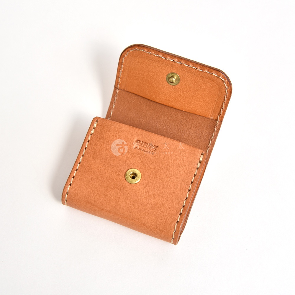 這是一款日本HERZ職人手作的棕色皮革零錢包，採用耐用的Stare皮革製成，表面刻有品牌LOGO。皮夾設計簡約精緻，中央配有黃銅金屬按扣，並以粗縫線手工縫製邊緣，呈現出溫潤且具質感的皮革工藝。