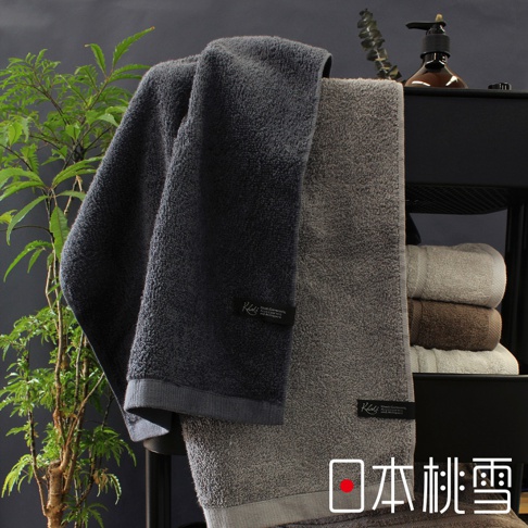 兩條厚實的男人大毛巾，一條為深灰色，另一條為淺灰色。毛巾質地蓬鬆，皆有品牌標籤，背景有綠植與其他顏色毛巾堆疊。