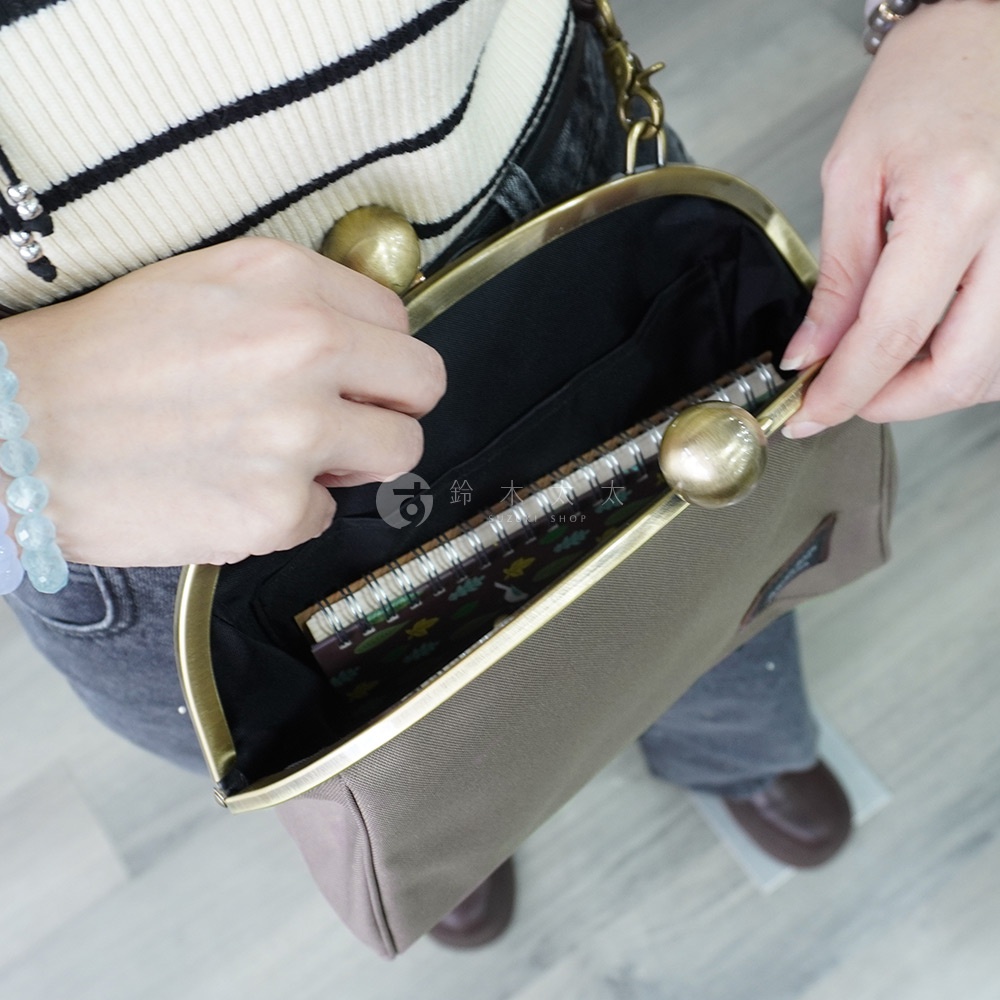 一位女性雙手正打開一個灰褐色帆布質料的側背包，包包上方有兩個古銅色大圓球金屬開釦，內側可見一本綠色封面筆記本，包包肩帶是粗獷鍊條式，使用者戴著一串淺藍色手鍊，穿著橫條紋毛衣與牛仔褲