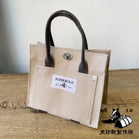 淺卡其色純棉帆布迷你托特包，配有深棕色提把和銀色旋轉鎖扣。包包前方有一個大口袋，上面貼有印有日文的白色標籤，標籤上有狗和袋子的圖案。