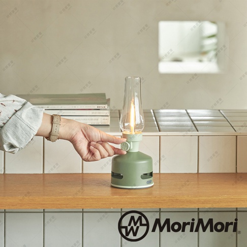 淺綠色煤油燈造型藍牙音響，玻璃燈罩內有LED燈，底部有網狀喇叭孔，一只戴著米色錶帶手錶的手正操作音響的旋鈕。