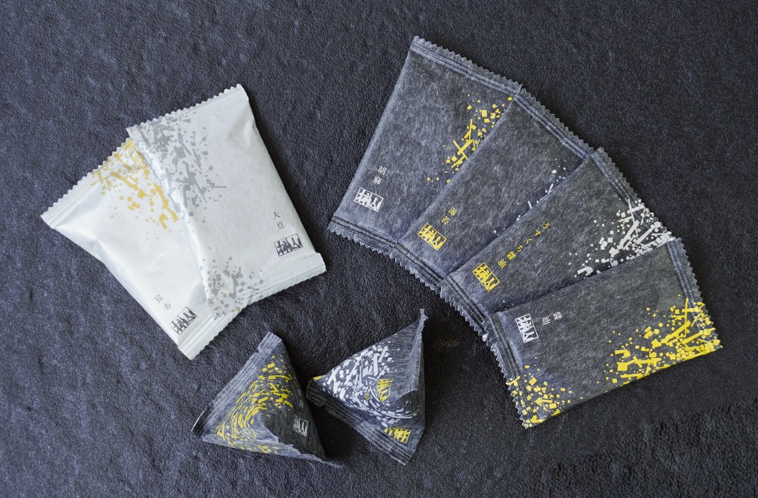 頂級米菓禮盒，包含兩包淺色調（白色與米色）的個別包裝，以及五包深灰色調的個別包裝，其中部分包裝為三角形。淺色包裝印有銀色與金色的抽象線條圖案，深灰色包裝則印有金黃色與白色的碎塊狀圖案，並有日文標示。
