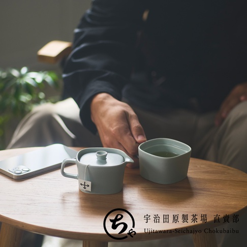 一位穿著深色衣服的人坐在木質圓桌旁，桌上擺放著宇治田原製茶場的淺綠色美濃燒一人用茶具組，包含一個帶蓋的小茶壺和一只馬克杯，旁邊還有一支智慧型手機，整體氛圍寧靜舒適。
