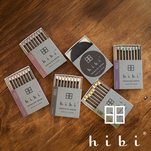 木質桌面上有五盒 hibi 香氛火柴以及一個圓形黑色燃燒墊。每盒火柴包裝上皆印有品牌標誌與產品編號，包含 001、002、003、004、005 等不同香氣款式，火柴棒頂端呈現不同色彩，整體設計簡約雅致，呈現日本工藝的質感。