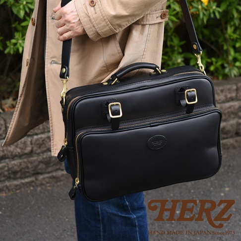 黑色皮革兩用商務公事包，配有金色搭扣和拉鍊，以及可拆卸肩帶。包身正面有兩個拉鍊隔層，中央有一個圓形壓花品牌標誌。