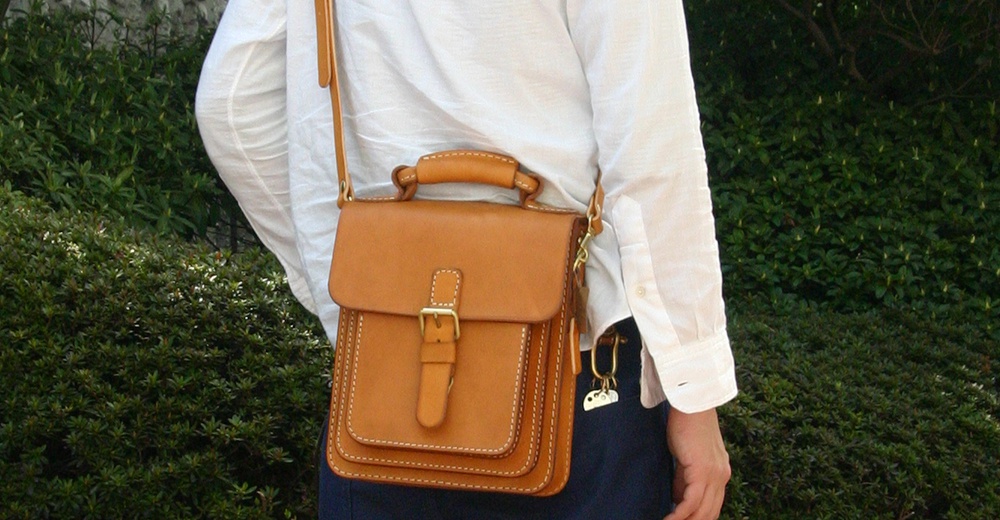 職人手作皮革單肩兩用小方包，淺棕色，有翻蓋及金屬扣，附有手提帶及斜肩帶，側邊有掛飾。