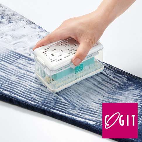 手持一個透明塑膠盒，內有藍綠色海綿和白色顆粒，盒蓋為白色帶有透氣孔，盒身上有「uchi more LAUNDRY」字樣，正置於沾有泡沫的藍色牛仔布上。