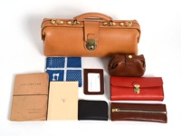 皮革製成的橫長形杜勒斯單肩包，搭配金屬鎖扣及提把。周圍擺放著棕色小零錢包、紅色長夾、棕色拉鍊皮夾、黑色拉鍊皮夾、深棕色卡夾、藍色白點圖案手帕，以及兩本不同顏色的筆記本。