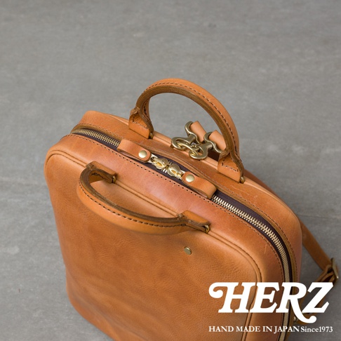 HERZ 品牌標誌，下方為 HAND MADE IN JAPAN Since1973 字樣，整體為一個棕褐色皮革後背包的特寫，包身帶有拉鍊和金屬鎖扣，採用近距離俯視角度拍攝。