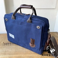 深藍色純綿帆布商務公事包，B4尺寸，配有棕色皮革提把和可拆卸肩帶，正面有金屬扣環和品牌標籤。