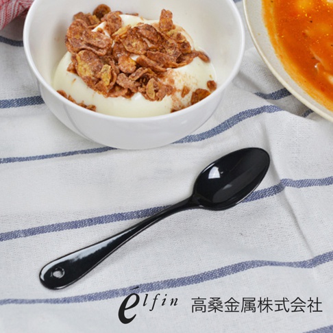 黑色琺瑯水果甜點匙，勺柄末端有圓孔，置於白色條紋布上，旁有一碗裝有優格及麥片，以及一盤橘色食物。