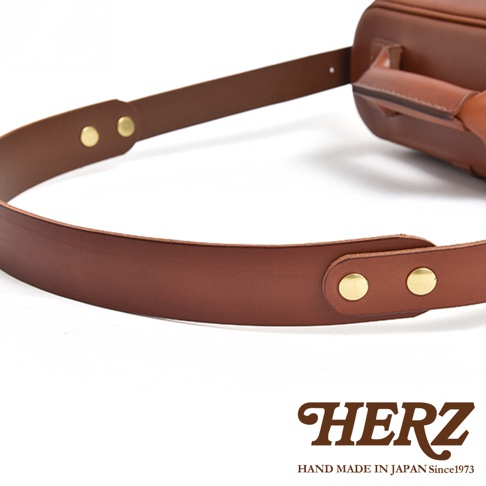 HERZ 品牌標誌，下方為 HAND MADE IN JAPAN Since1973 字樣，圖片主體為皮革相機包的棕色皮革背帶，特寫細節可見鉚釘。