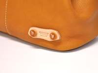 近距離特寫，展示職人手作皮革托特包的局部，包身呈現溫潤的焦糖棕色，材質為柔軟的真皮，表面帶有細緻的紋理。包側裝飾有品牌皮革標籤，以雙扣環固定，縫線細膩。