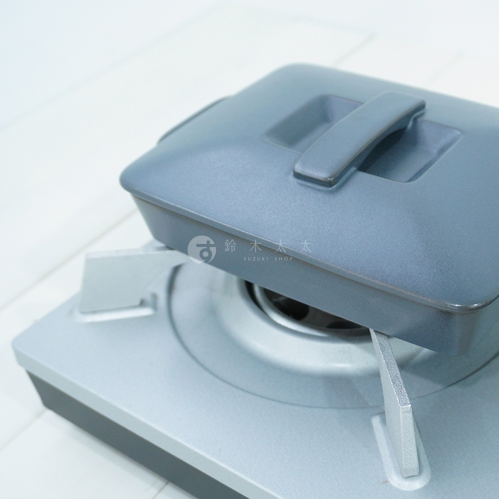 灰藍色陶製萬用烤盤，附有同色系的方形提把蓋子，置於銀灰色爐架上，呈現簡約的廚房烹飪情境。