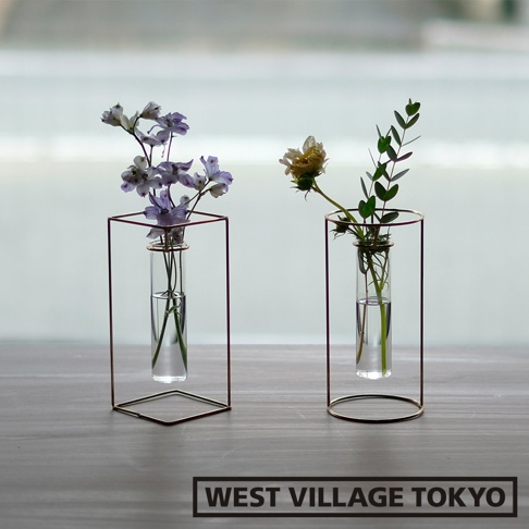 兩個黃銅四角玻璃花器，分別插著紫色小花和乾燥花束，置於木質桌面，背景為模糊的窗景。