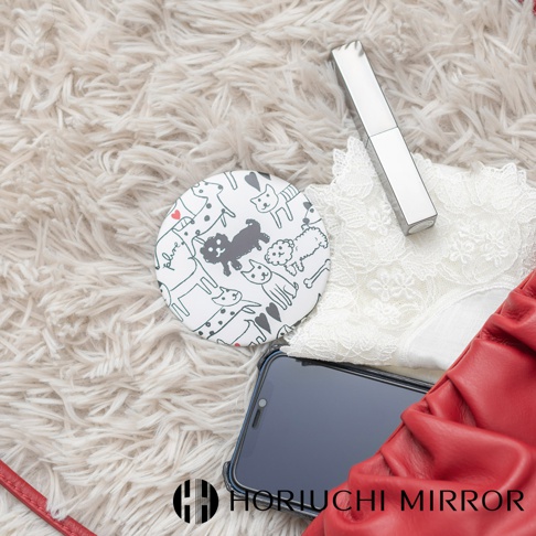 白色圓形化妝鏡，鏡面印有黑白簡筆畫風格的狗狗圖案，其中一隻為黑色。鏡子旁有銀色唇膏、白色蕾絲布料、黑色手機和紅色皮革包。